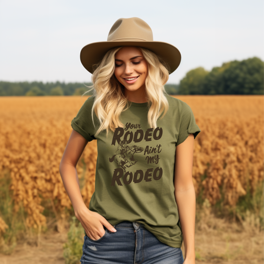 Woman wearing a green t-shirt with 'Your Rodeo Ain't My Rodeo' text and a hat, standing in a field.